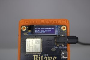 A close-up view of a Bitaxe Bitcoin miner in a custom orange "SOLO SATOSHI" case, displaying setup instructions on its OLED screen. The screen reads "Connect to the configurati... Wi-Fi (for setup): Bitaxe_0469," indicating the device is in WiFi setup mode.