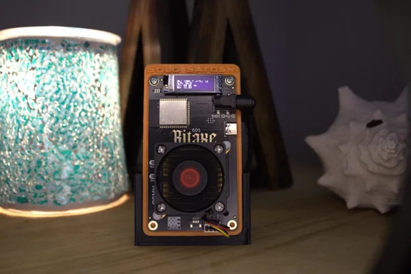 Bitaxe Gamma solo Bitcoin miner on desk stand with OLED display lit and red 40 mm fan beside turquoise lamp and shell décor.