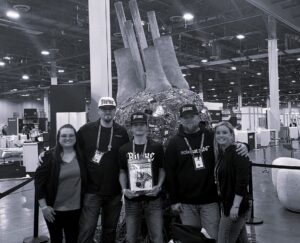 Solo Satoshi team at Bitcoin conference, posing in front of a circuit board art installation, proudly representing the Bitaxe brand.