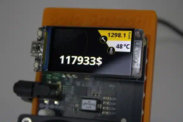 Close-up of NerdAxe Gamma solo Bitcoin miner’s LILYGO OLED display showing 1.298 TH/s hash-rate, 48 °C ASIC temperature and live BTC price on an orange Solo Satoshi stand.