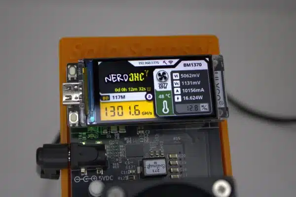 Close-up of NerdAxe Gamma Bitcoin solo miner displaying 1.30 GH/s hash-rate, 48 °C chip temperature and 12.8 W/TH efficiency on the OLED screen