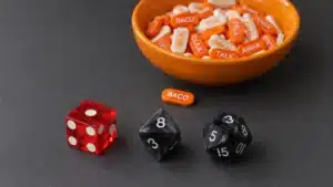 Entropia v2 seed phrase entropy tablets in an orange bowl beside a red six-sided casino die, a black eight-sided die, and a black sixteen-sided die on a dark work mat