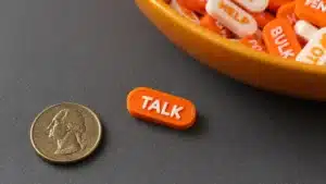 Single Entropia v2 seed phrase tablet reading TALK face-up on a dark work mat beside a coin, with a bowl of mixed orange and white tablets in the background